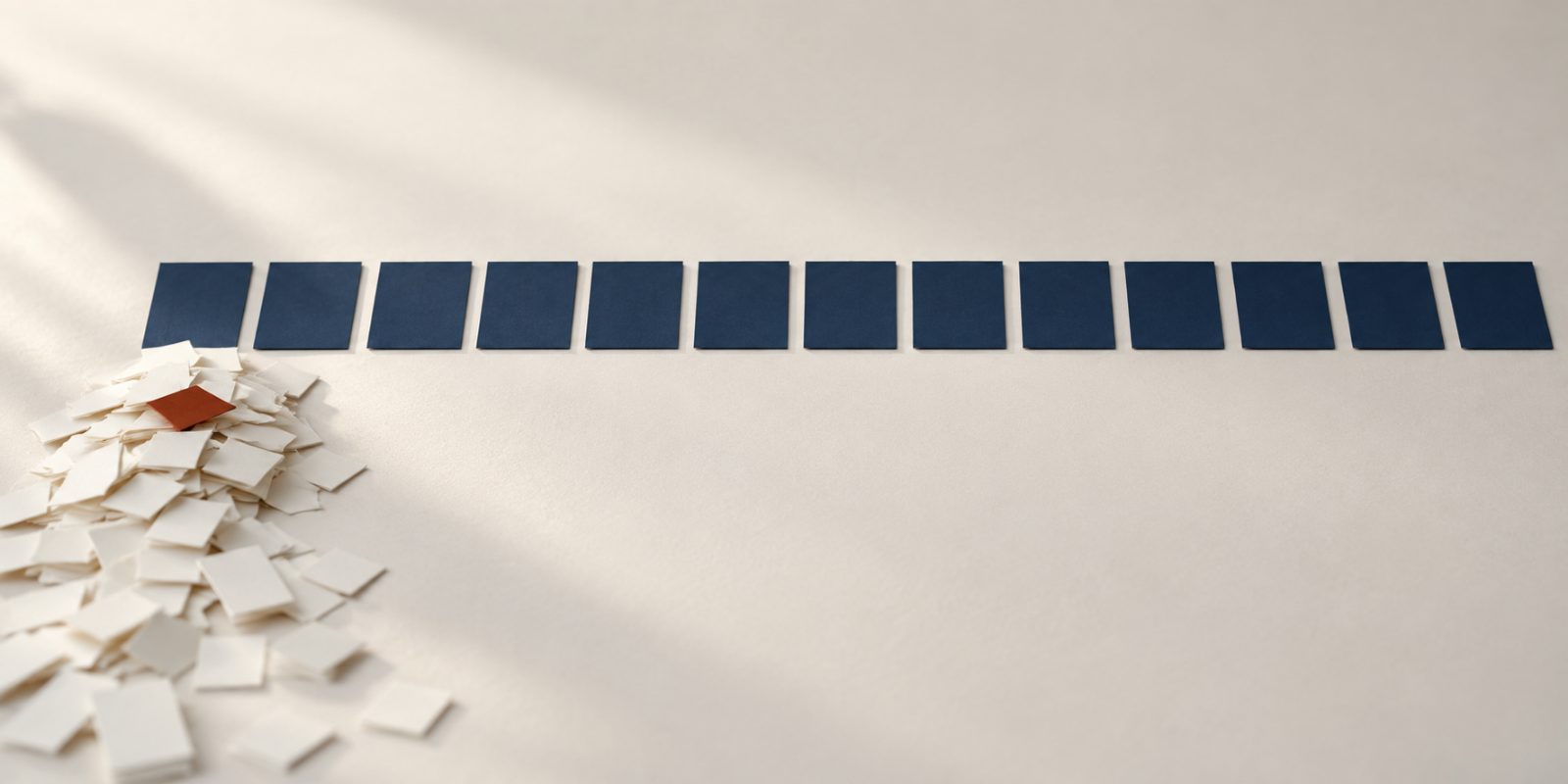 A photographic still life in the JobCraftly brand colours. A row of thirteen identical blank deep-navy rectangular cards lies evenly spaced across a pale warm off-white surface in soft side light. Beneath the first card at the left end of the row a heap of small blank pale paper slips has piled up and spills towards the front of the frame; one slip near the top of the heap is deep rust red. The surface under the other cards is empty.