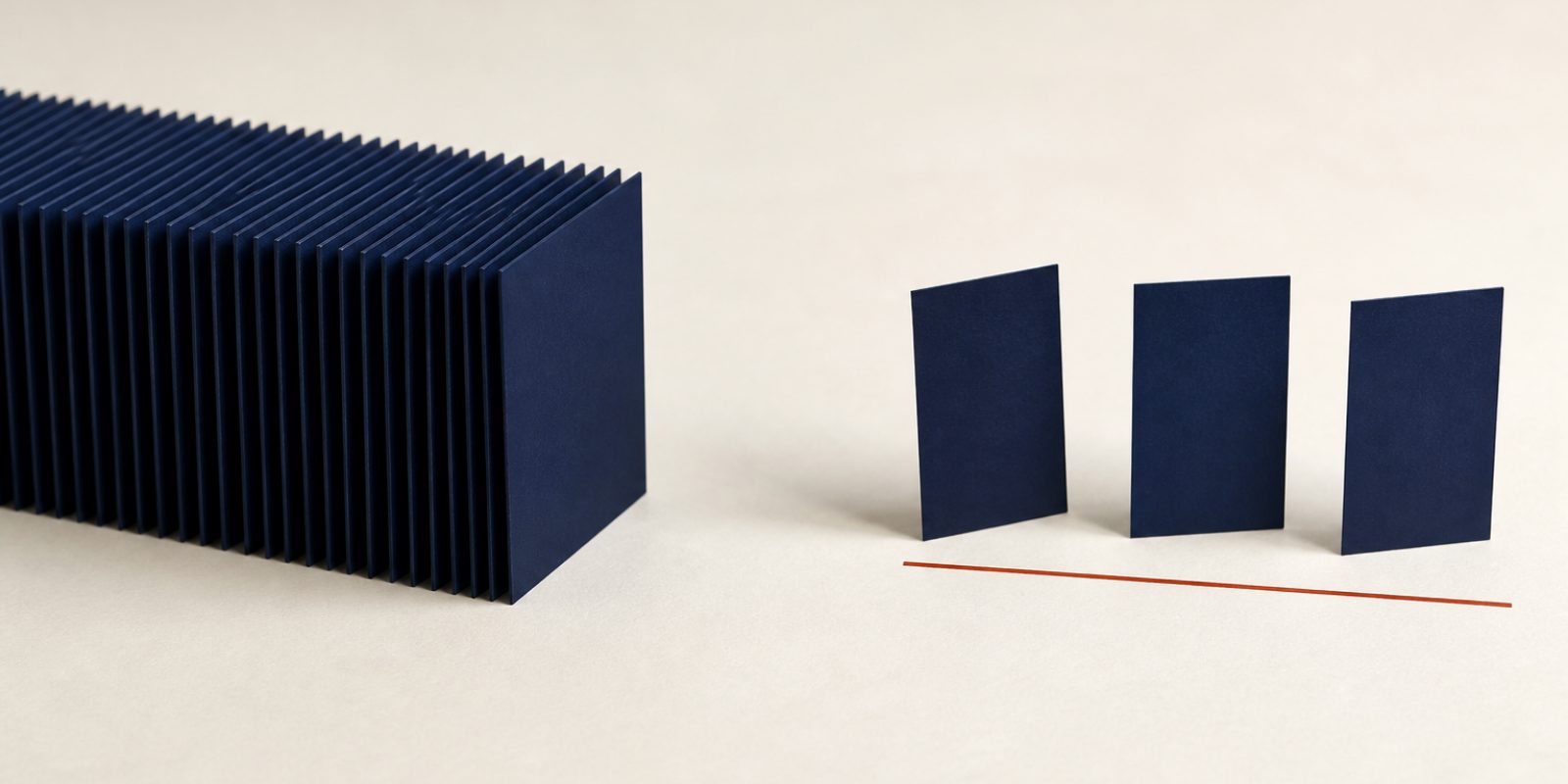 A photographic still life in the JobCraftly brand colours. On the left, a long block of many identical blank deep-navy cards stands upright on a pale warm off-white surface, packed so tightly together that only their edges show. On the right, across an empty gap, three of the same blank navy cards stand well apart from each other, each leaning at a slightly different angle, with a thin deep rust-red line running along the surface in front of them.