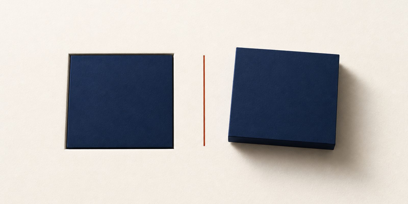Two blank deep-navy squares of the same size on a pale warm off-white surface in soft light. The left one is set flush into a precisely cut recess, its top level with the surface. The right one rests loose on top, rotated slightly off square, its thickness visible along the bottom edge and a soft shadow falling away to the right. A short thin deep rust-red line stands vertically in the gap between them.