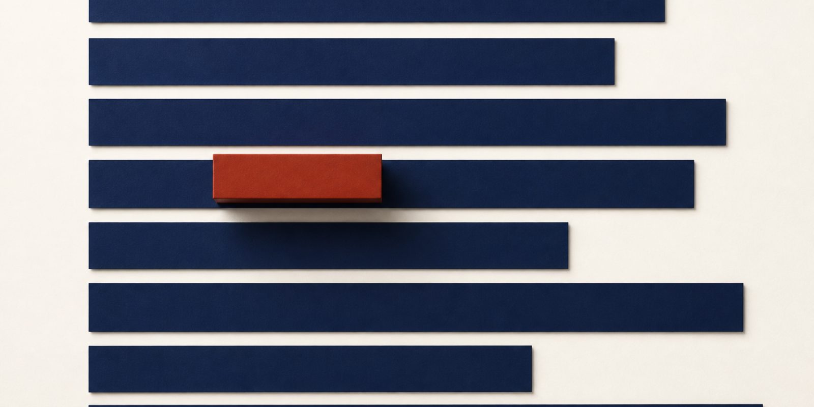Blank deep-navy paper strips of different lengths lying flat one below another on a pale warm off-white surface, each starting at the same left edge and ending at a different point across the frame. The top strip is cut in half by the top of the frame and only the edge of the lowest one is visible at the bottom. A solid deep rust-red block rests on the fourth strip down, raised above it and casting a soft shadow across the gap and the strip below.